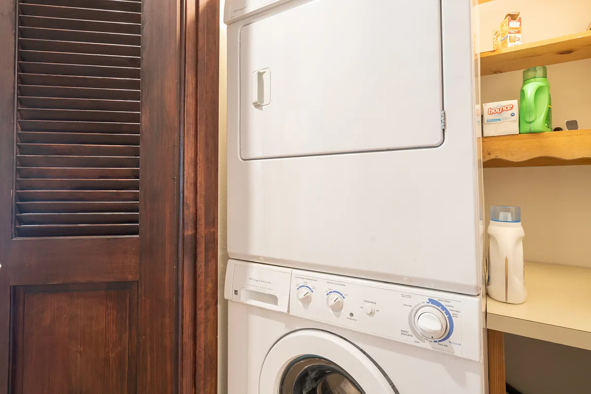 This image features a stacked washer and dryer set within a tidy laundry area, complete with shelves stocked with laundry supplies and a wooden door nearby. The setup conveys convenience and practicality for guests managing everyday needs.