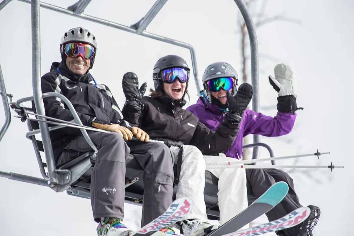 Three skiers ride a chairlift over a snowy landscape, outfitted in ski gear and surrounded by stunning mountain scenery. The camaraderie and anticipation of a ski day are palpable, emphasizing the proximity and appeal of the local slopes.