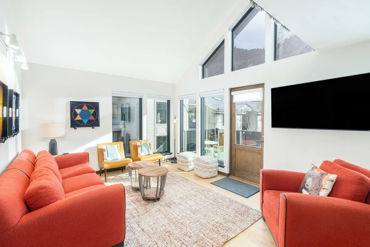 A cozy living room setup with red sofas, expansive windows revealing beautiful outdoor scenery, a wooden door, and colorful artwork. The arrangement encourages relaxation, socializing, and memorable family activities.