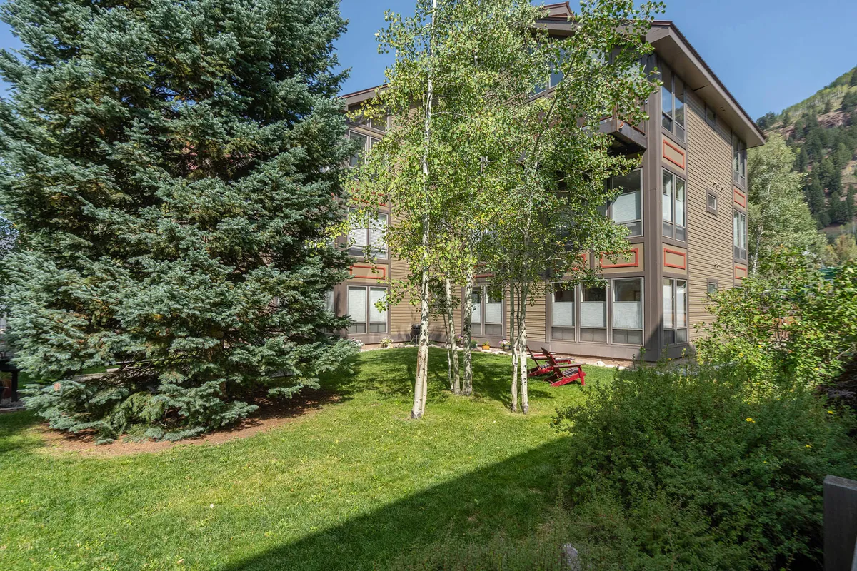 The image features the lodge’s facade nestled among green pine and aspen trees, with red chairs set in a grassy garden area. The tranquil outdoor space suggests opportunities for relaxation, play, and socializing just steps from the building.