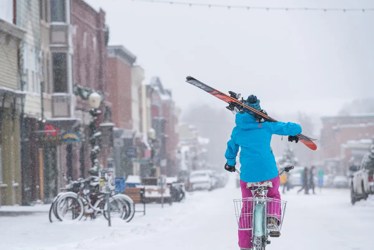 A lively street scene shows a person bicycling with skis in tow down a snow-covered street lined by colorful buildings. The vibrant atmosphere captures the blend of biking, skiing, and town exploration that defines Telluride’s active lifestyle.