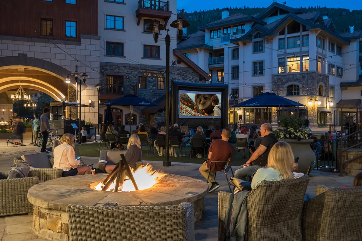 An inviting outdoor communal space featuring a fire pit, a projector screen for outdoor movies, comfortable seating, and surrounding buildings, set against an evening sky. People are gathered, enjoying a relaxed and social atmosphere.