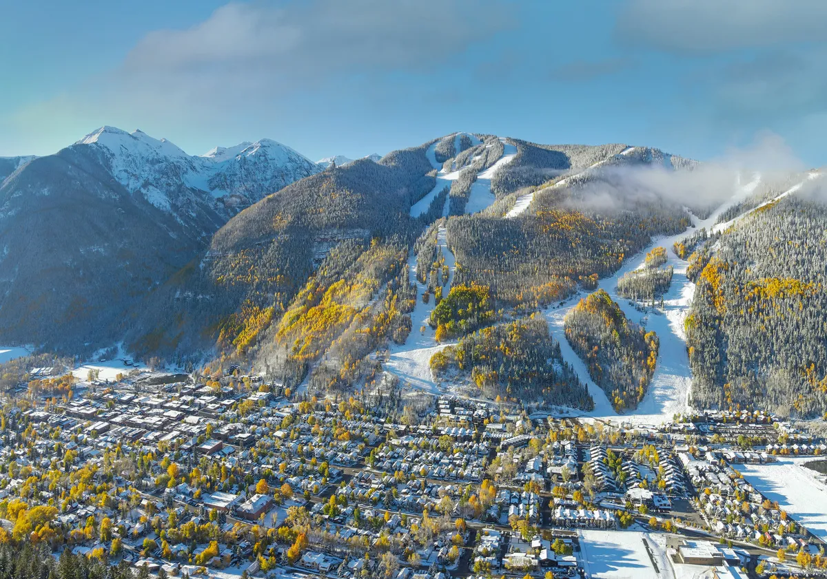 This scenic shot captures snow-capped mountains, ski slopes, colorful autumn trees, and the quaint town nestled below. The scene hints at both winter and summer adventures—skiing, hiking, and exploring local charm—with a sense of excitement and possibility.
