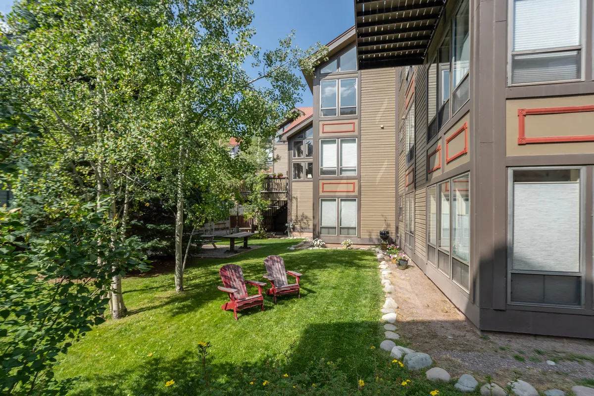 This exterior image showcases the community’s grassy, fenced-in yard with red Adirondack chairs placed on the lawn, the building’s facade, and surrounding trees. The relaxed outdoor setting invites residents to enjoy the communal yard for picnics, reading, and socializing with neighbors.