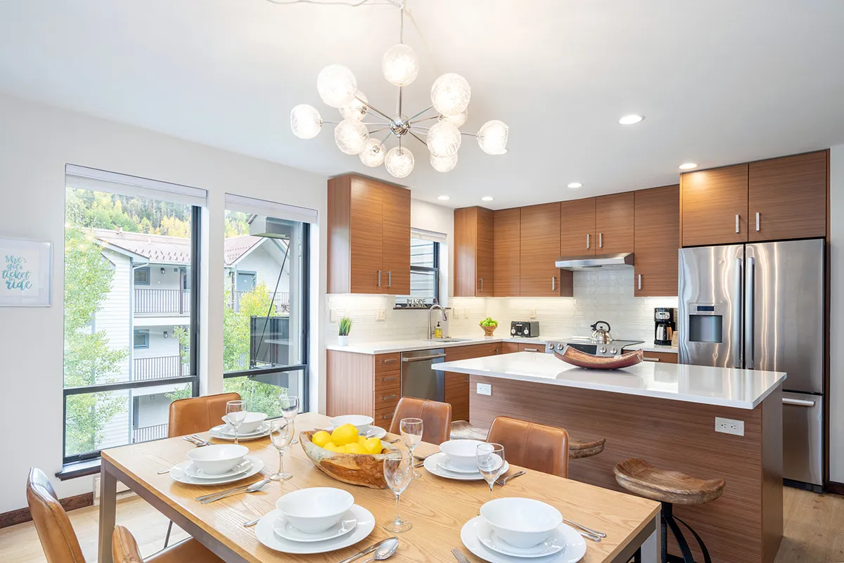 A spacious and inviting dining area is shown, with a modern chandelier above the dining table, a kitchen island nearby, and large windows letting in ample natural light. The layout is designed for family meals, games, or gatherings.