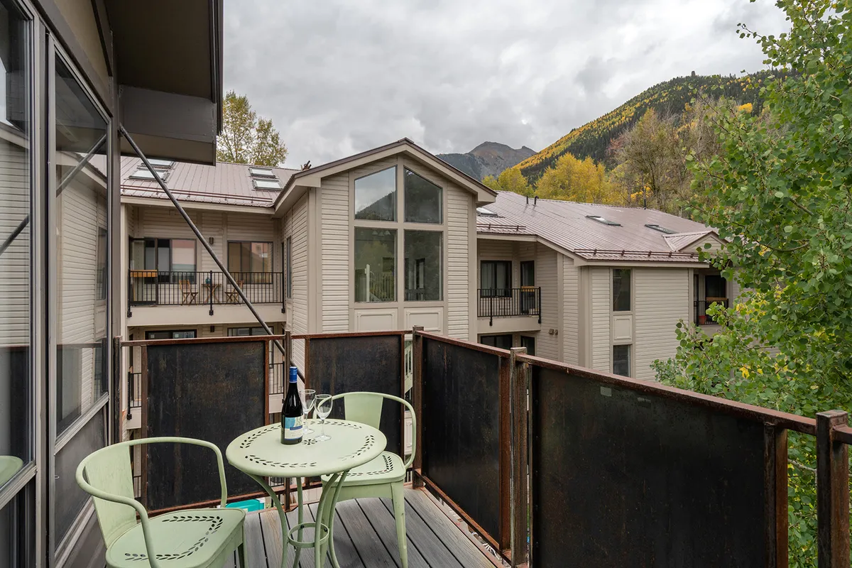 A tranquil private balcony scene with a small green table, two chairs, a bottle of wine, and sweeping mountain views framed by trees. This space is ideal for casual conversation or soaking up nature’s beauty.