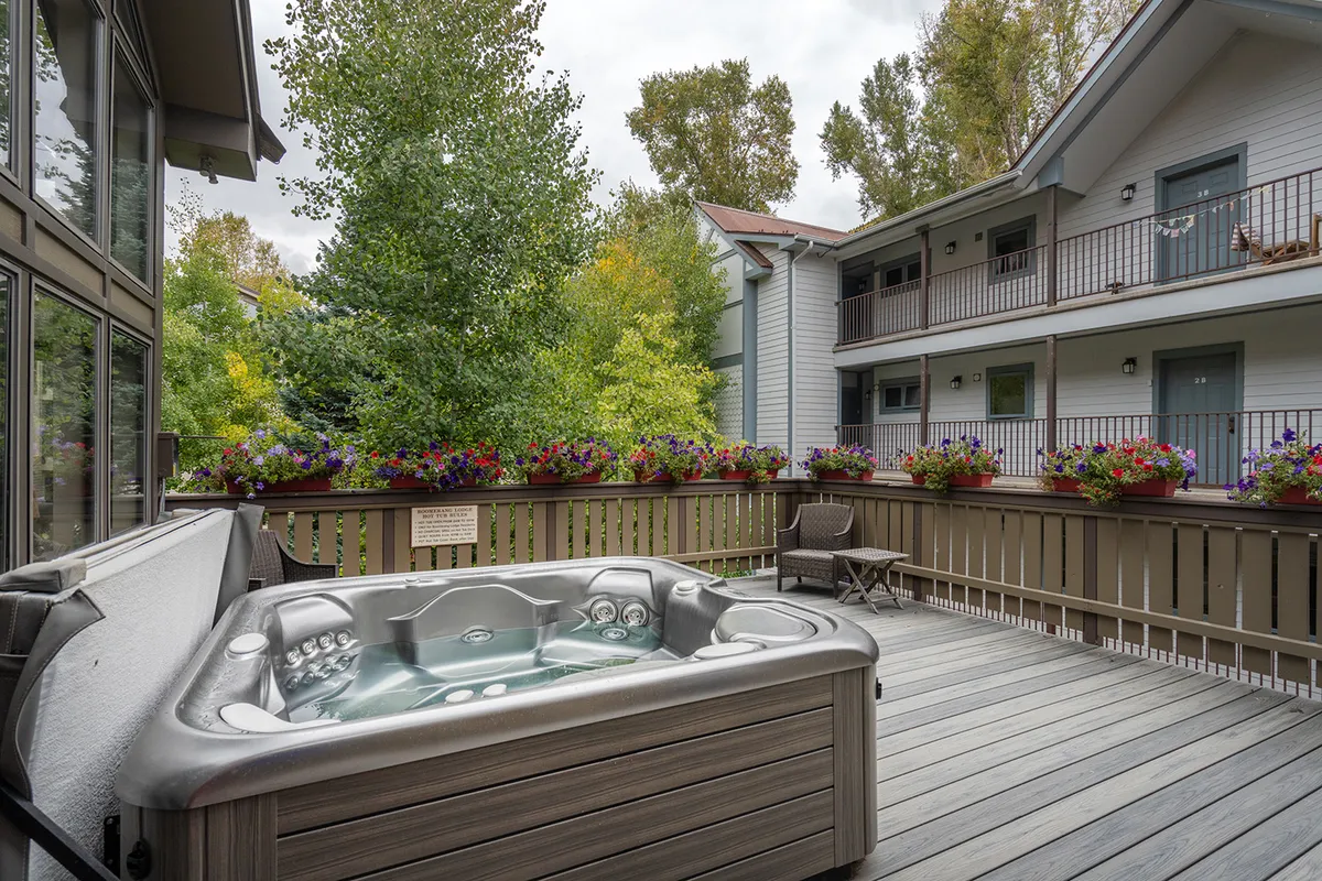 A tranquil outdoor deck features a communal hot tub surrounded by decorative flower pots and wooden decking, with greenery in the background. The space is designed for relaxation and social gatherings, appealing to residents after a day on the slopes or during summer evenings.