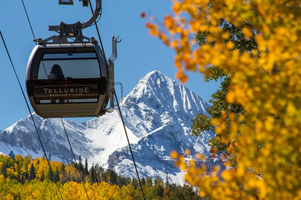 A gondola glides over vibrant autumn foliage and snow-capped peaks, capturing the excitement of seasonal change and accessibility to mountain trails. The gondola symbolizes easy, scenic access to both skiing and hiking opportunities.