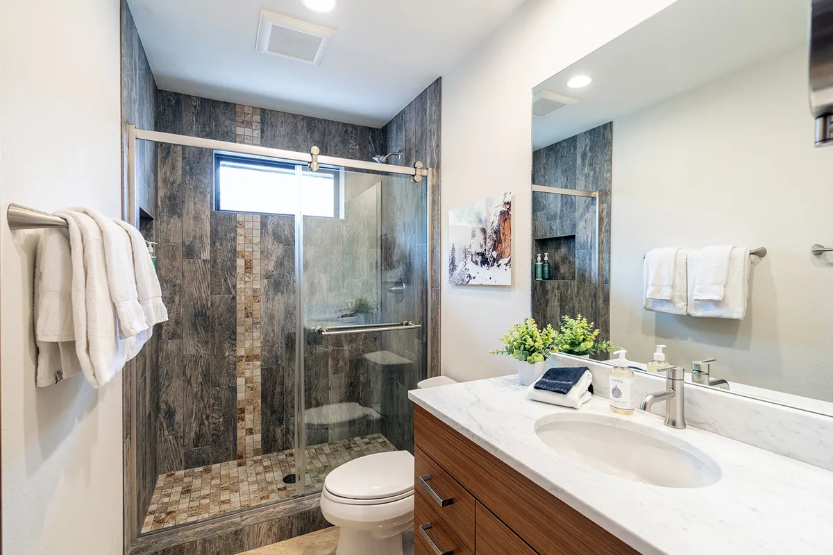 A modern, tranquil bathroom featuring a glass-enclosed shower, marble countertop, wood cabinetry, and soothing artwork. The space looks spa-like and comfortable, ideal for self-care routines or a relaxing wind-down at the end of the day.