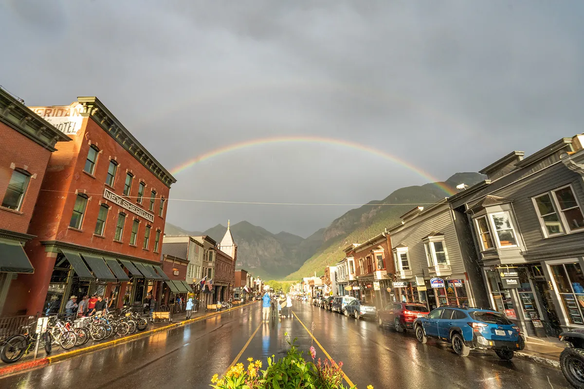 A vibrant street scene in Telluride featuring historic buildings, a glowing rainbow overhead, a wet street alive with the reflections of recent rain, people strolling, bicycles parked, and an overall lively, welcoming atmosphere.