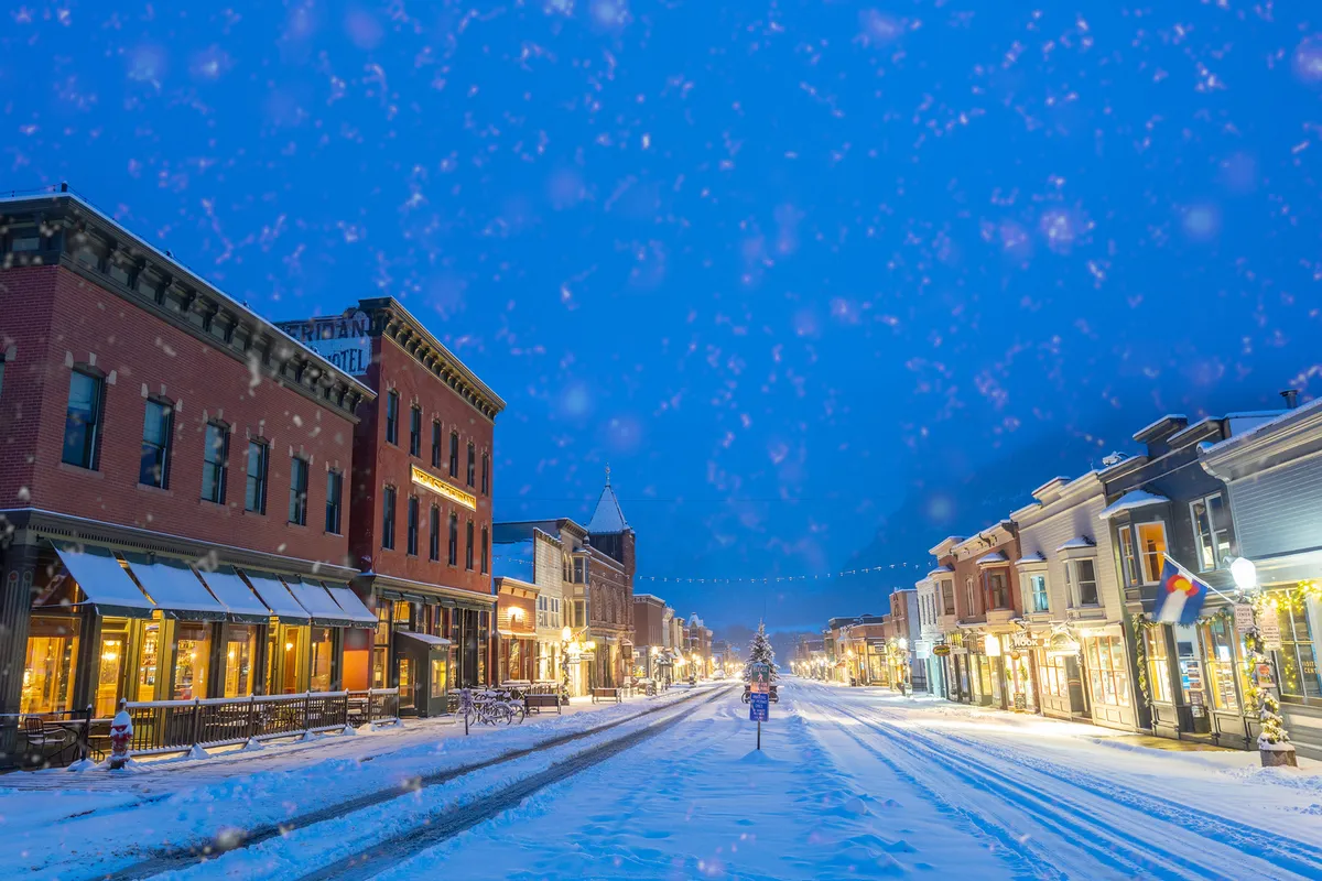 A picturesque snow-covered street in downtown Telluride, featuring decorated storefronts and a glowing Christmas tree that illuminate the festive winter atmosphere. The scene invites exploration of nearby boutiques and acclaimed restaurants.