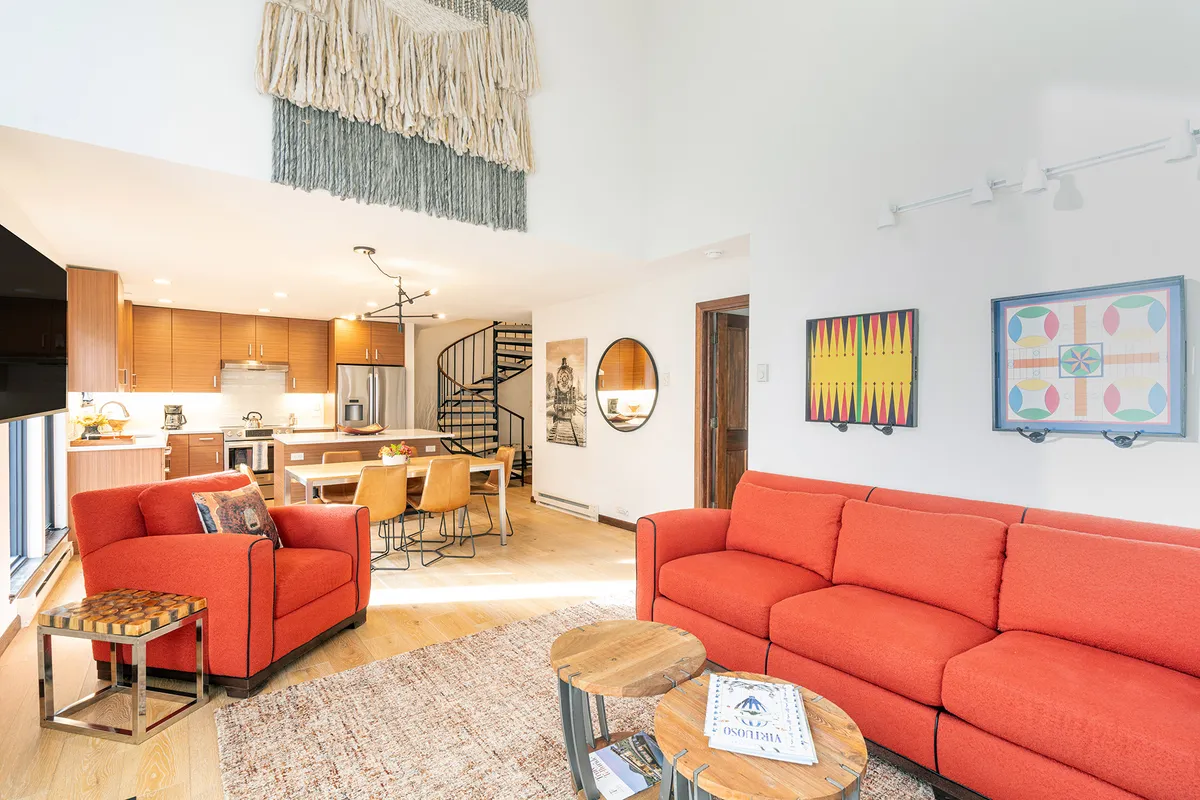 A sunlit, contemporary living space featuring a vibrant red sectional sofa, a striking spiral staircase, artisanal textile wall art, and an inviting dining area—perfect for gathering and entertaining.
