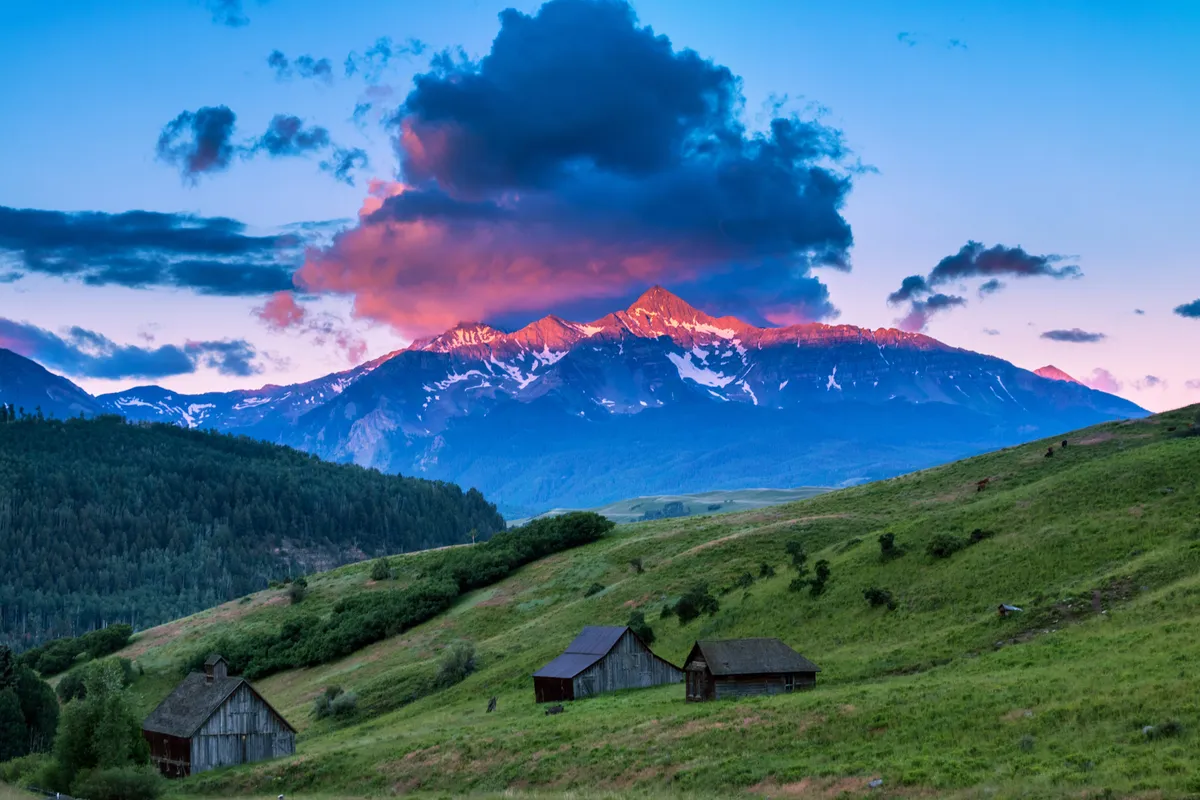 A wide-angle view of snow-capped mountains, glowing clouds at sunset, green rolling hills, and rustic wooden cabins. The peaceful landscape evokes a sense of escape and harmony with nature.