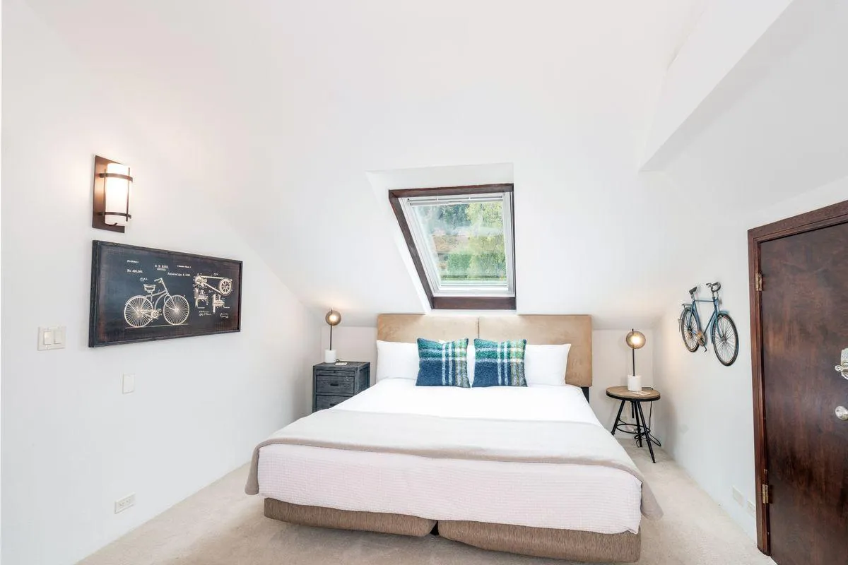 A peaceful bedroom with a neatly made bed, soft lighting, a window offering natural light, and tasteful artwork. The space feels restful and inviting, suitable as either a queen or additional guest room, perfect for quiet reading or relaxation.