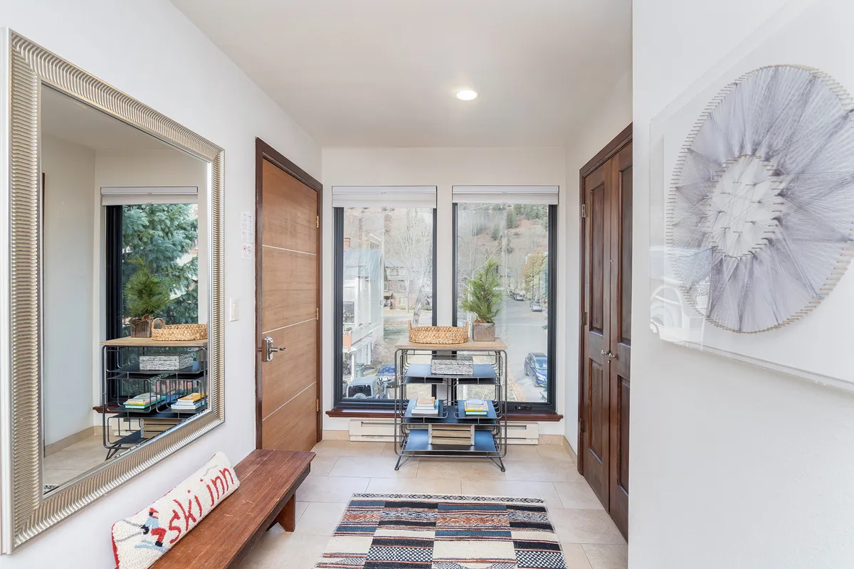 This image captures the entry foyer, featuring a bench, a large mirror, and a floor-to-ceiling window that fills the space with inviting natural light. Subtle decorative art and a shelving unit create a sense of welcome and warmth.
